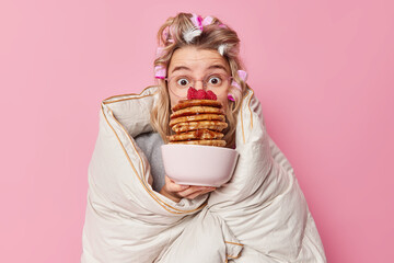 Impressed young European woman with hair rollers wrapped in duvet holds bowl of appetizing pancakes...