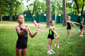 Girl doing exercise with clubs on rhythmic gymnastics training with other trainees outdoors in summer in sports camp