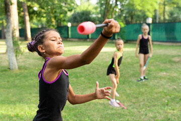 Girl doing exercise with clubs on rhythmic gymnastics training with other trainees outdoors in...