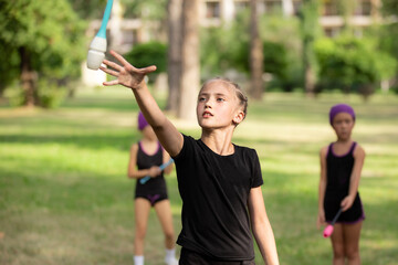 Obraz premium Girl doing exercise with clubs on rhythmic gymnastics training with other trainees outdoors in summer in sports camp