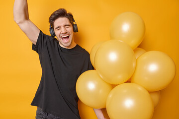 Cheerful adult man exclaims from joy keeps arm raised up sings favorite song dressed in casual black t shirt wears stereo headphones on ears holds bunch of inflated balloons isolated over yellow wall