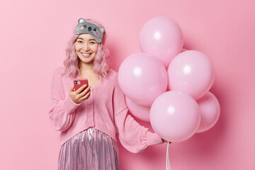 Obraz na płótnie Canvas Positive Asian woman with pink dyed hair uses mobile phone gets messages of congratulation on her birthday wears festive clothes holds bunch of inflated balloons isolated over pink background