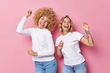 Cheerful young friendly women have good time dance and foolish around laugh gladfully dressed in casual clothes isolated over pink background feel energetic take break listen favorite music.
