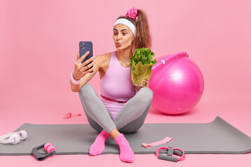 Obraz na płótnie Canvas Fitness and workout concept. Lovely sporty European woman dressed in sportswear holds lettuce salad and makes selfie via smartphone keeps to diet does gymnastics at home uses sport equipment