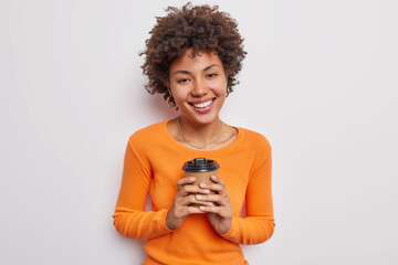 Positive young beautiful woman with dark curly hair holds disposable paper cup of coffee enjoys drinking caffeine beverage wears casual orange jumper isolated over white background poses indoors - Powered by Adobe