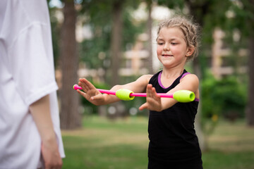 Girl trainee showing exercise with clubs to her coach on rhythmic gymnastics training outdoors in...