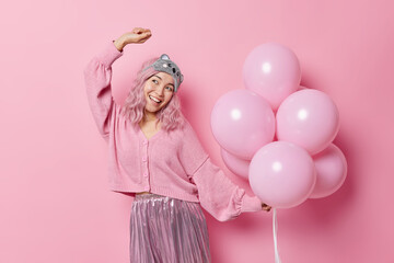 Happy Asian woman with dyed hair good mood wears blindfold on forehead jumper and skirt dances carefree has fun on party holds bunch of inflated helium balloons isolated over pink background