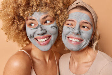 Two happy friendly young woman stand closely to each other appy clay facial masks smile toothily...