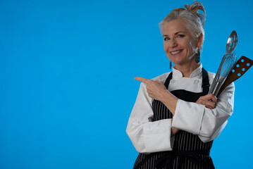 Portrait professional female chef in uniform looking and pointing at copy space on solid blue...