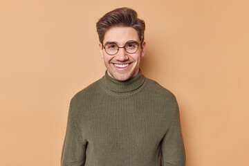 Portrait of positive guy with dark hair smiles happily wears round transparent eyeglasses and...