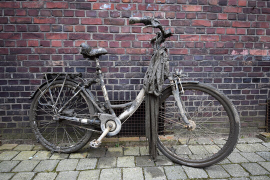 Bicycle covered in mud and silt, dredged out of canal or river, standing or parked against orange brick wall