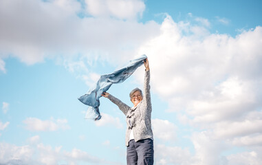 Happy mature elderly woman outdoors waving blue scarf in the wind. Smiling caucasian elderly woman with short hair wearing glasses enjoying freedom and vacation