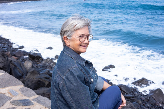 Portrait of white-haired smiling senior woman sitting on the beach wearing eyeglasses and denim jacket. Attractive 70 years old woman enjoying good time and vacation