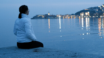 A girl in a white jacket looks at the evening sea in blue tones