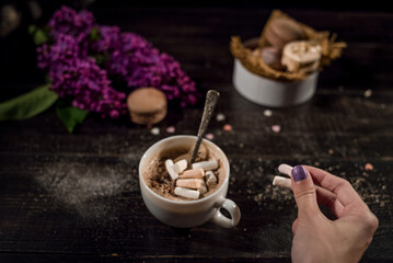 Hot chocolate (cocoa) in a glass mug with two tubes for drinking.There are some macarons on a linen napkin on the wood table with some lilac