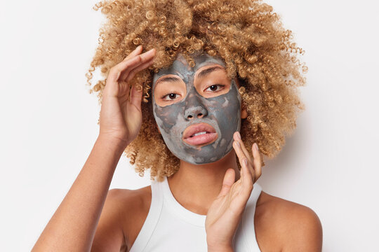 Photo of serious charming curly woman touches face gently applies clay facial mask to look beautiful wears casual t shirt poses against white background shows bare shoulders. Skin care concept