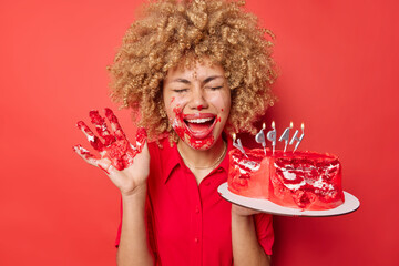 Cheerful curly haired woman foolishes around dirty with cream eats tasty sweet cake with hands...