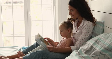 Young caring foster mom sit on cozy bed hug little adopted daughter read story with kid engage preschooler in learning activity. Happy Latin mom look at pictures in colorful book with small child girl