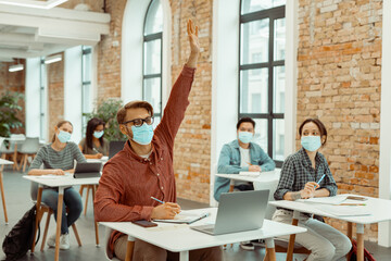Male student in facial mask raising hand for answer