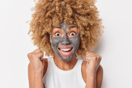 Headshot of happy curly haired woman looks excitedly clenches fists from joy applies facial clay mask for skin treatment wears casual t shirt isolated over white background. Skin care concept