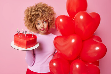 Upset bored European woman holds delicous heart shaped cake and bunch of red helium balloons has spoiled makeup wears casual jumper isolated over pink background. People and holidays concept