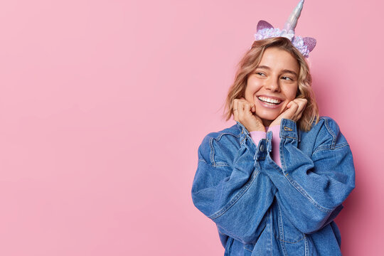 Indoor shot of positive young European woman keeps hands under chin wears unicorn headband and denim jacket smiles toothily has curious expression isolated over pink background copy space for text