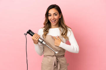 Young woman using hand blender over isolated pink background with surprise facial expression