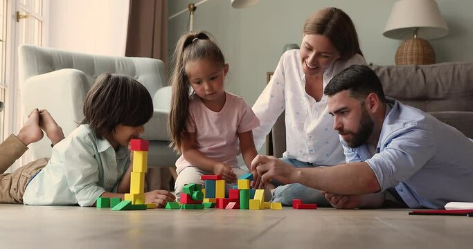 Young couple play wooden colored cubes with cute siblings. Full family sit on floor in living room spend weekend leisure at modern house enjoy game and communication. Children development, fun concept