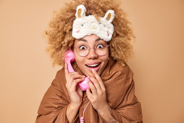 Cheerful surprised woman wrapped in blanket holds retro handset near ear has conversation before going to sleep reacts on amazing news wears sleepmask and spectacles isolated over brown background.