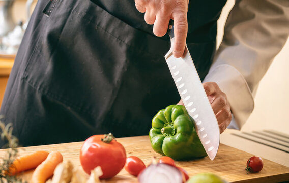 Chef Cut A Green Pepper With A Knife On A Chop Board, Close Up. Fresh Vegetable On Kitchen Table.