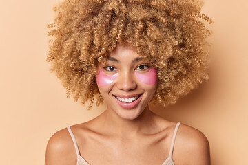 Close up shot of positive glad curly haired woman applies pink hydrogel patches under eyes smiles pleasantly wears t shirt stands bare shoulders isolated over beige background uses cosmetic product