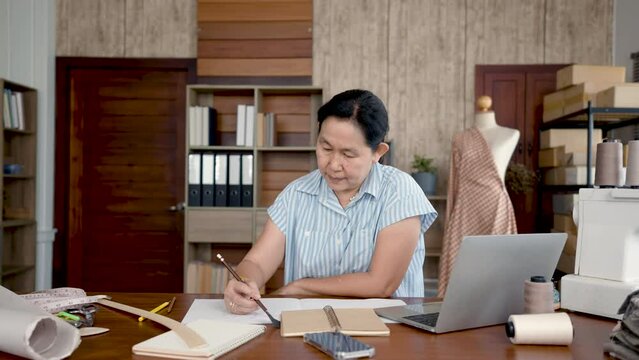 4K 50fps, Professional small business owner partner in clothing design,An Asian woman artisan sits and draws designs for customized outfits that customers have ordered on a board placed on the table.