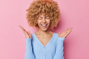 Happy emotional woman with curly bushy hair keeps palms raised laughs joyfully keeps eyes closed...