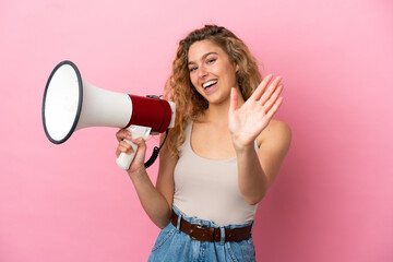 Young blonde woman isolated on pink background holding a megaphone and saluting with hand with...