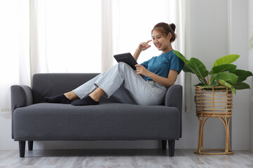 Fototapeta na wymiar Cheerful Asian young woman lifestyle while sitting on sofa at home using tablet and having fun.