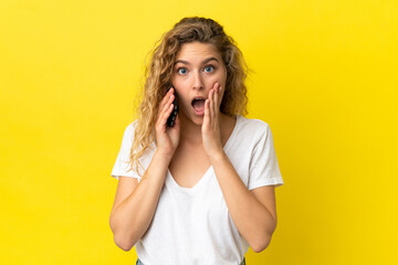 Young blonde woman using mobile phone isolated on yellow background with surprise and shocked facial expression