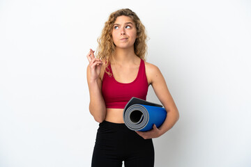 Young sport blonde woman going to yoga classes while holding a mat isolated on white background with fingers crossing and wishing the best