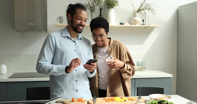 Young Black spouses couple in love chat talk share information on smartphones using wifi internet distracted from cooking food. Loving husband show picture on phone screen to smiling wife at kitchen