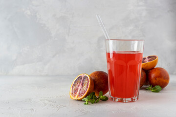 A tall glass of red orange juice on a light table. Red oranges and mint leaves in the background. Light background, space for text