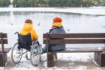 Caucasian woman in a wheelchair and her friend are sitting by the lake with ducks in winter.