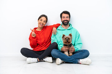 Young caucasian couple sitting on the floor with their pet isolated on white background smiling and showing victory sign