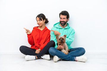 Young caucasian couple sitting on the floor with their pet isolated on white background pointing finger to the side and presenting a product