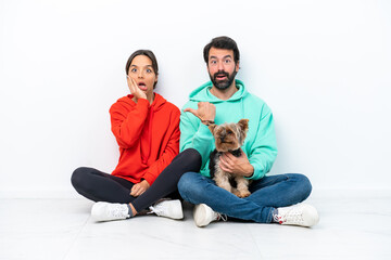 Young caucasian couple sitting on the floor with their pet isolated on white background pointing finger to the side and presenting a product