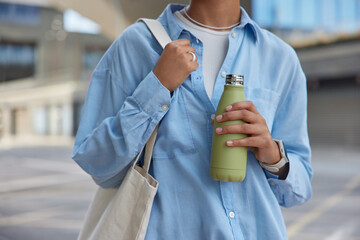 Cropped shot of unknown faceless woman wears blue shirt carries fabric bag holds bottle with fresh...