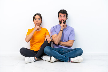 Young caucasian couple sitting on the floor isolated on white background showing a sign of closing mouth and silence gesture