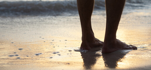 Male feet in the water on the sand beach, sunset reflection on the wet sand. Barefoot man stands in the water on the beach.