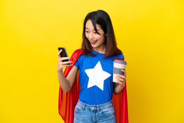 Super hero Vietnamese woman isolated  on yellow background holding coffee to take away and a mobile