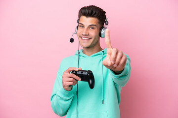 Young handsome caucasian man playing with a video game controller isolated on pink background showing and lifting a finger © luismolinero