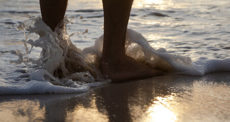 Male feet in the water on the sand beach, sunset reflection on the wet sand. Barefoot man stands in the water on the beach.