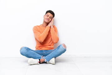 Young man sitting on the floor isolated on white background making sleep gesture in dorable expression